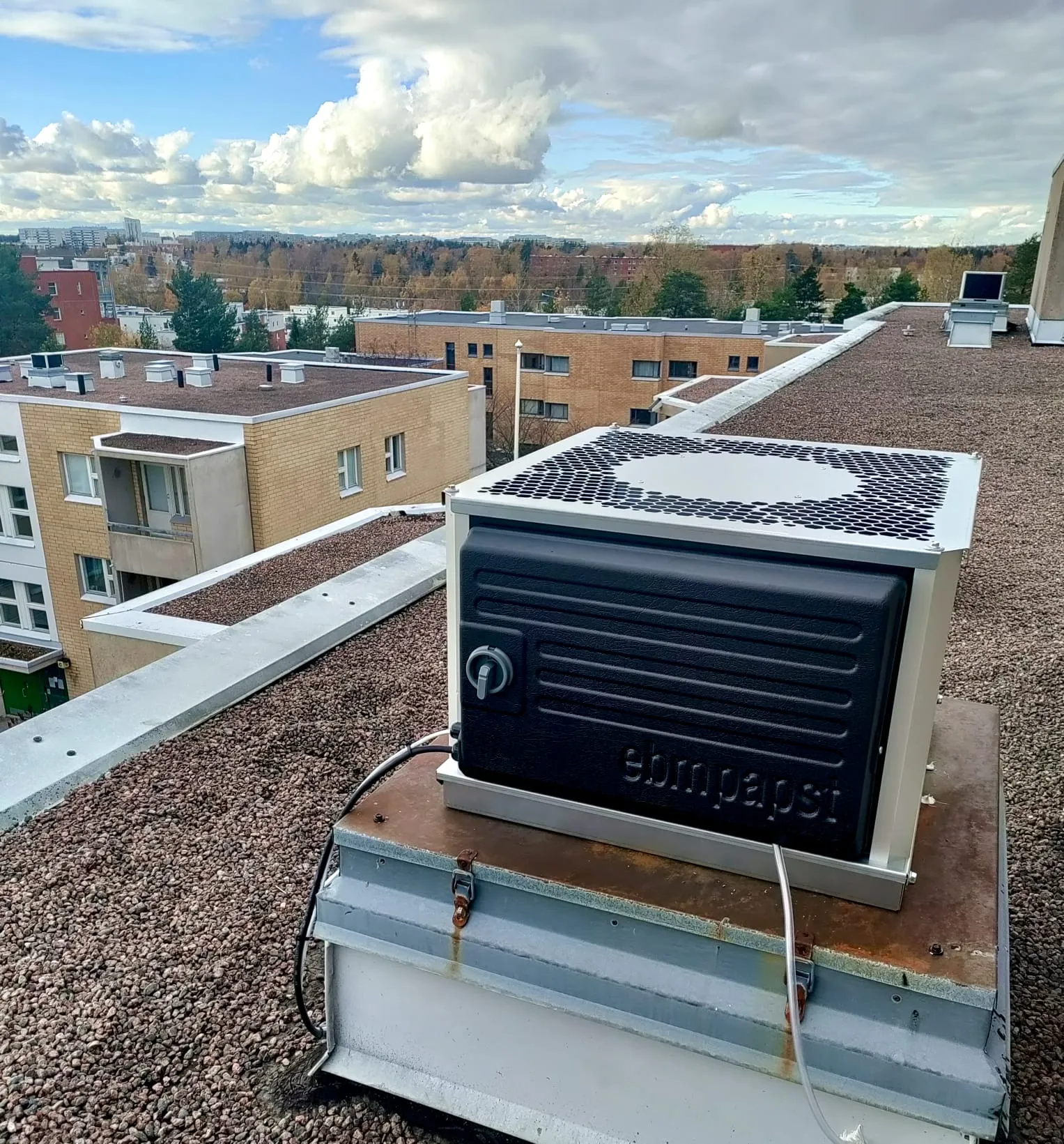 Kattoasennuslaite, jossa on musta kotelo ja valkoinen ritilä, sijaitsee kivikattoisella rakennuksella, taustalla rakennuksia ja taivas.