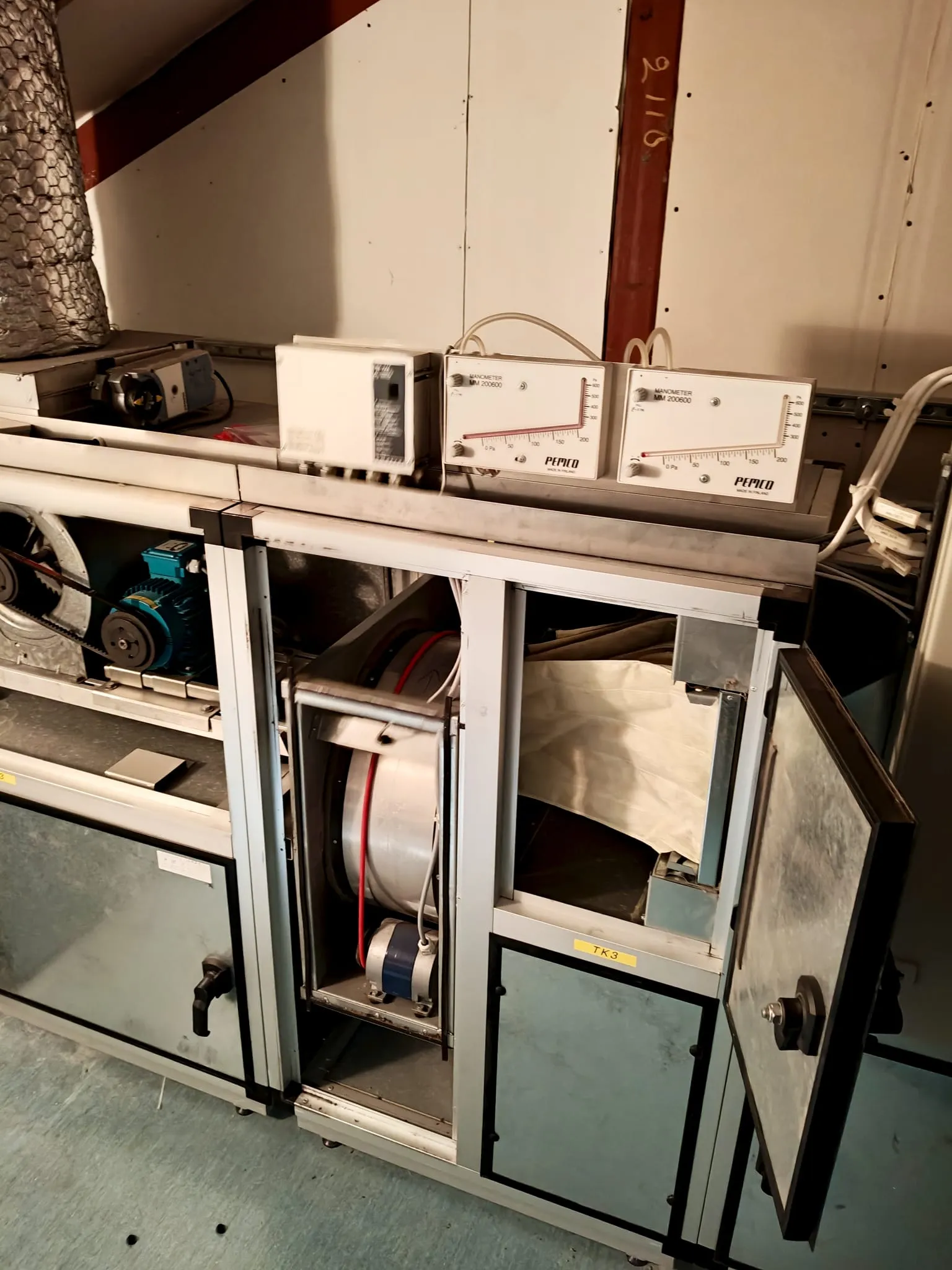 Ilmastointilaite, jossa on avoin ovi, kaksi manometriä yläosassa, harmaa metallinen runko ja epätasainen valaistus.