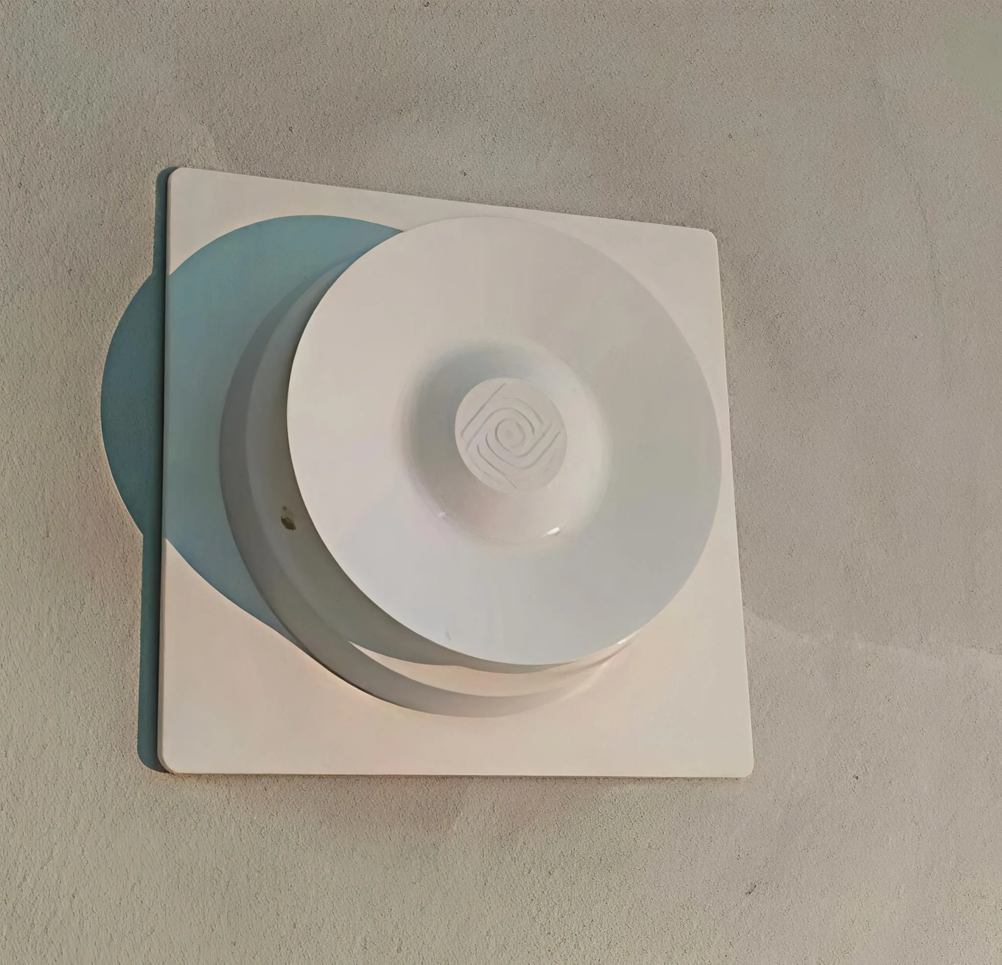 Valkoinen, pyöreä ilmanvaihtoventtiili neliönmuotoisella pohjalla, asennettu vaaleanharmaaseen seinään, varjo vasemmalla.