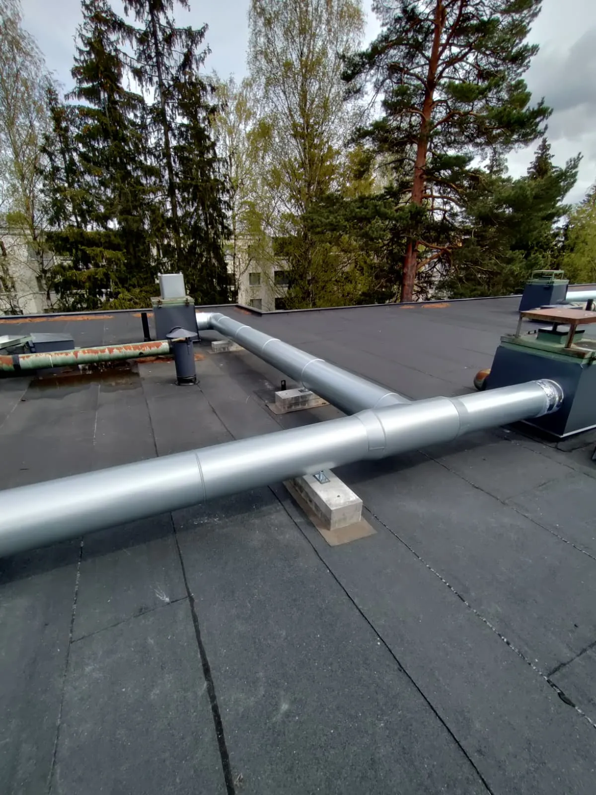 Kaksi hopeanväristä putkea kulkee mustalla katolla, taustalla puita ja rakennuksia, valoisa sää.