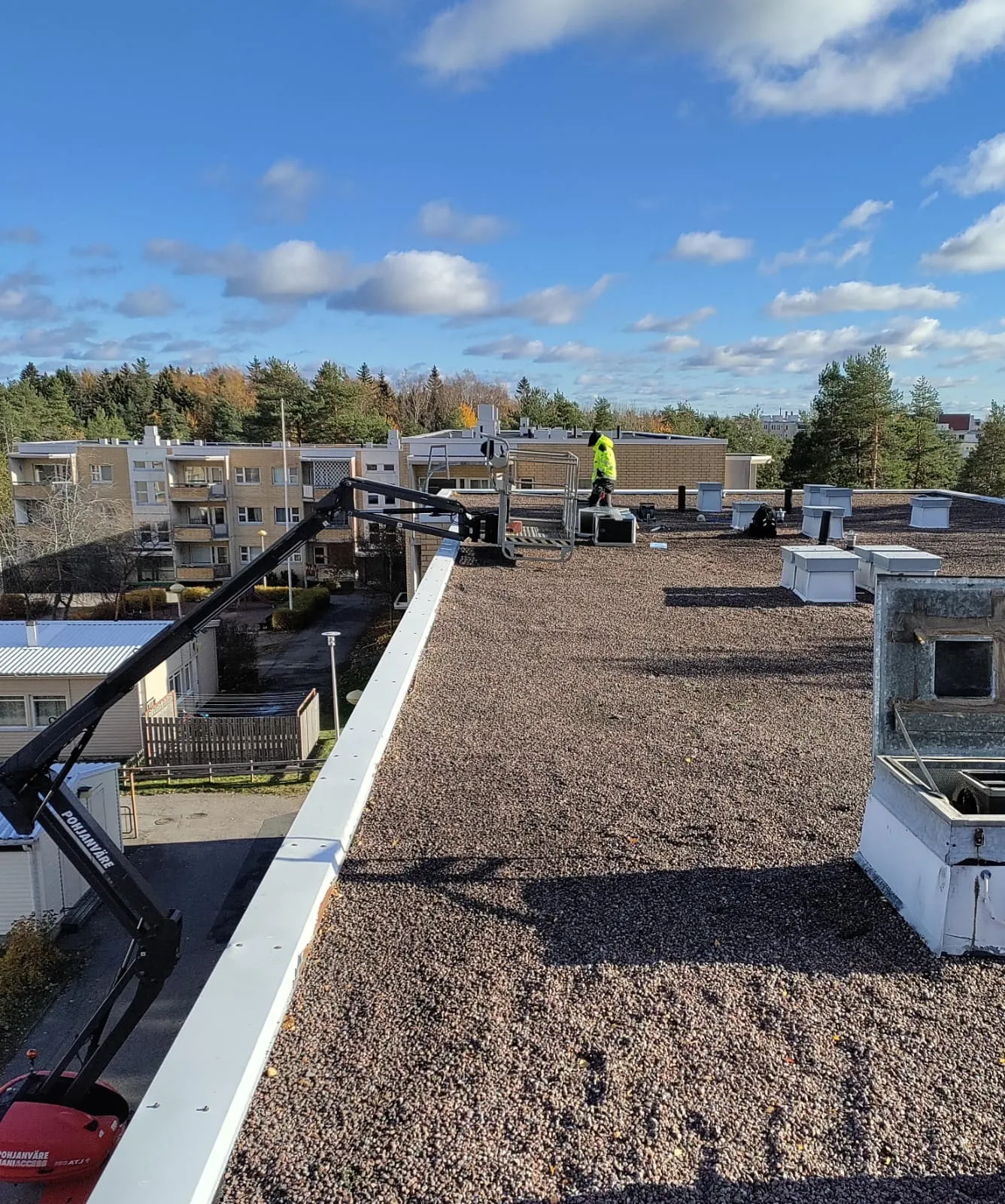 Näkymä katolta, jossa on sorapäällyste, valkoinen reunus ja nosturi. Työntekijä seisoo katolla keltaisessa turvaliivissä. Taustalla näkyy rakennuksia ja puita sinisen taivaan alla.