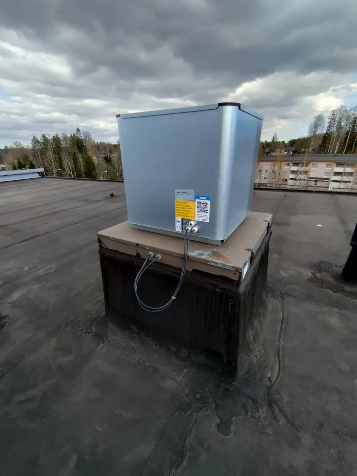 Harmaa laatikko katolla, jossa on kaksi johtoa ja tarra, taustalla pilvinen taivas ja rakennukset.