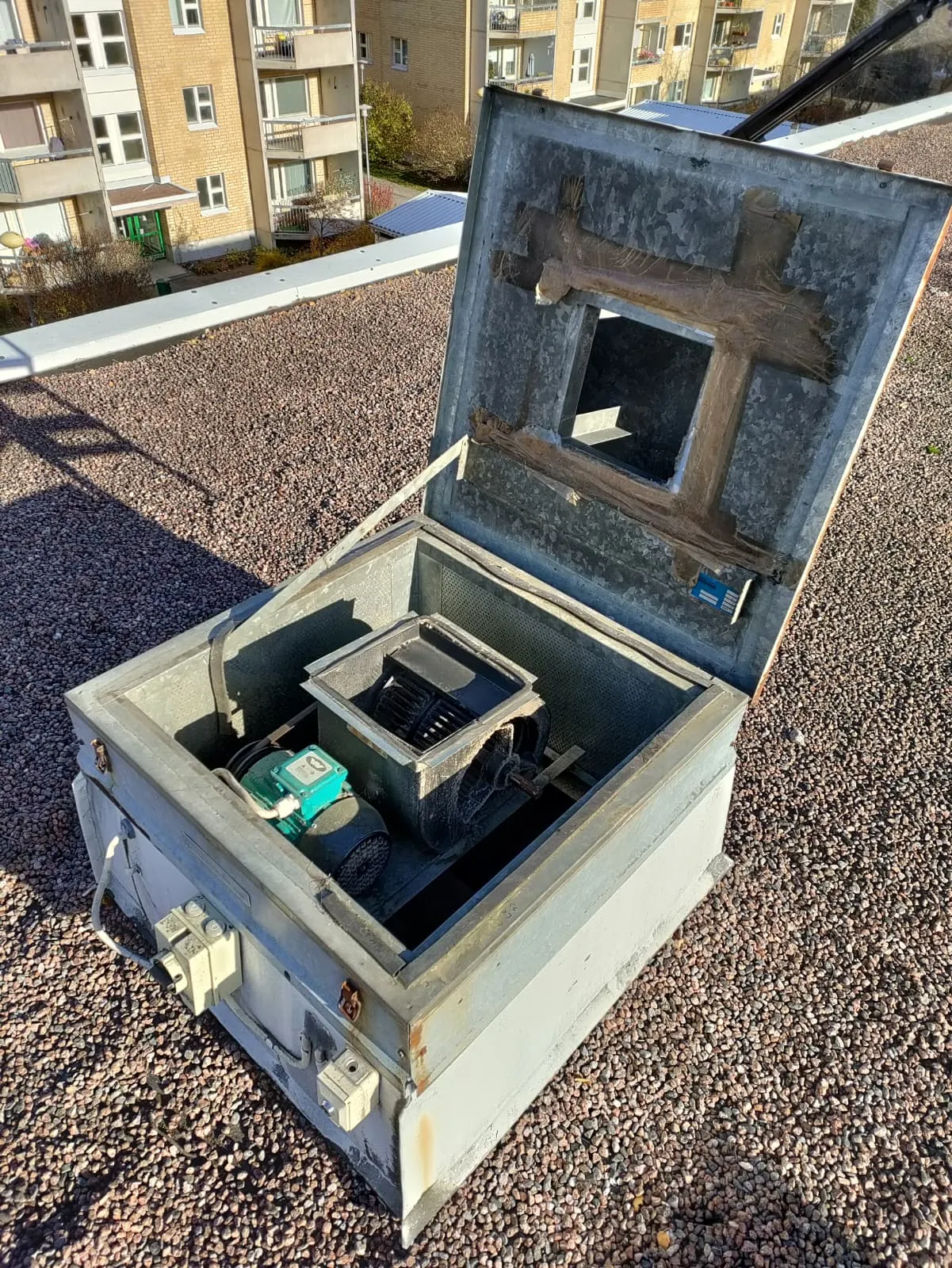 Avoin metallinen ilmastointilaite kivikattoisella rakennuksella, jossa näkyy moottori ja tuuletin.