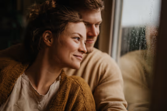 A couple embracing and looking out a window on a rainy day, sharing a warm moment together.