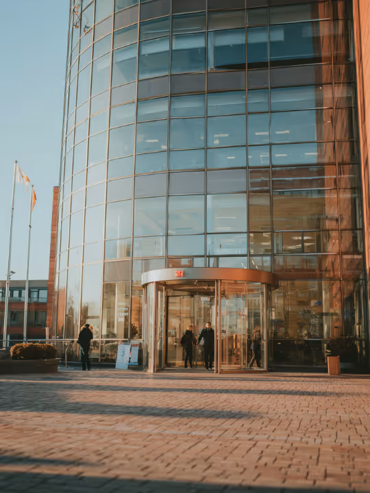 Modern office building with large curved glass facade and revolving door at entrance, people walking nearby.