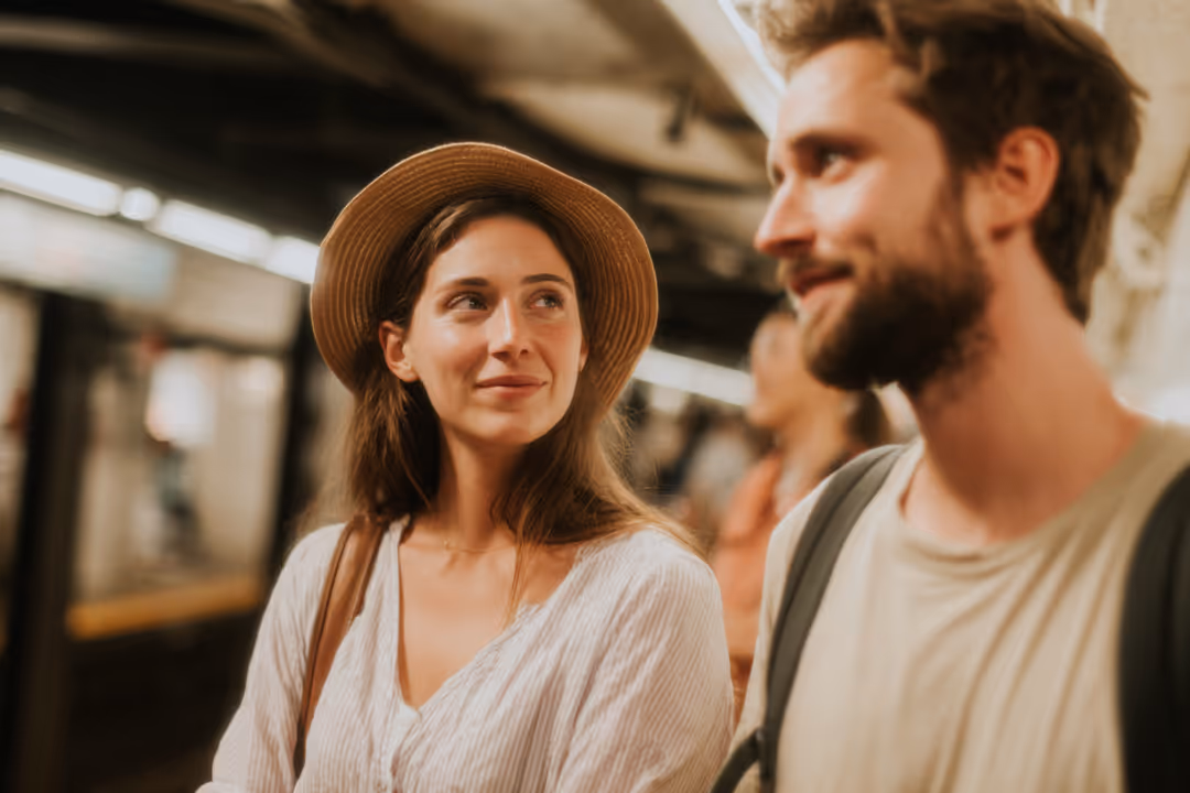 Smiling young woman wearing a hat looking at a bearded man with a backpack in a subway station.