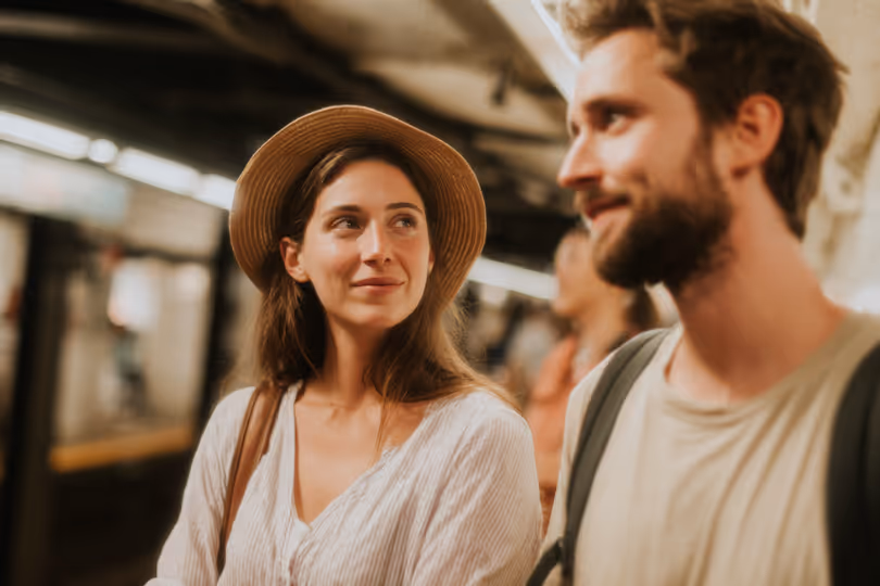 Young woman wearing a hat looking at a smiling man with a beard in a subway station.