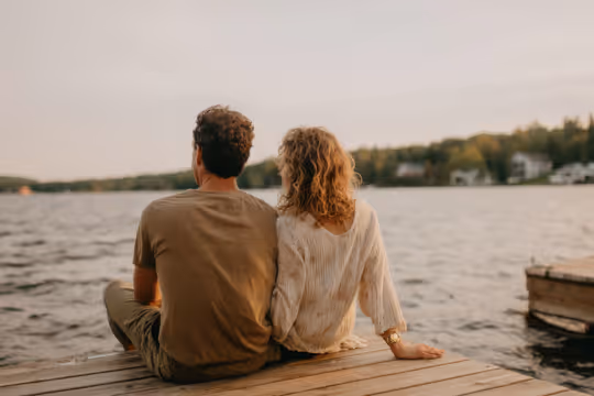 Couple sitting on a wooden dock facing a calm lake during sunset.