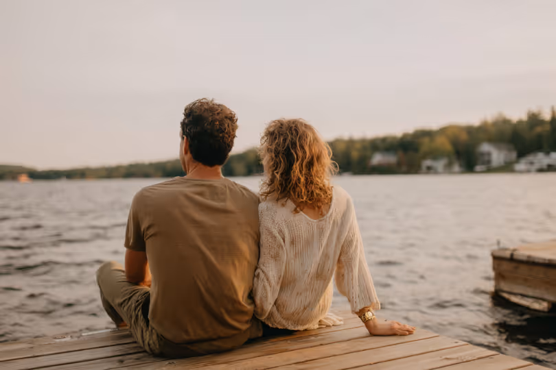 Couple sitting side by side on a wooden dock overlooking a calm lake during sunset.