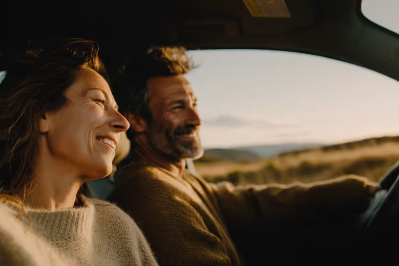 Middle-aged couple smiling and enjoying a car ride during sunset with a scenic landscape outside.