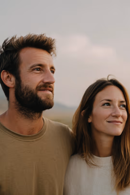 Man and woman standing close together outdoors, looking into the distance with soft smiles.