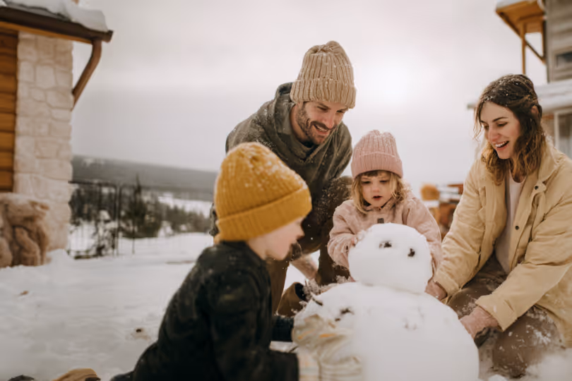 Family of two adults and two children building a snowman outdoors in winter.