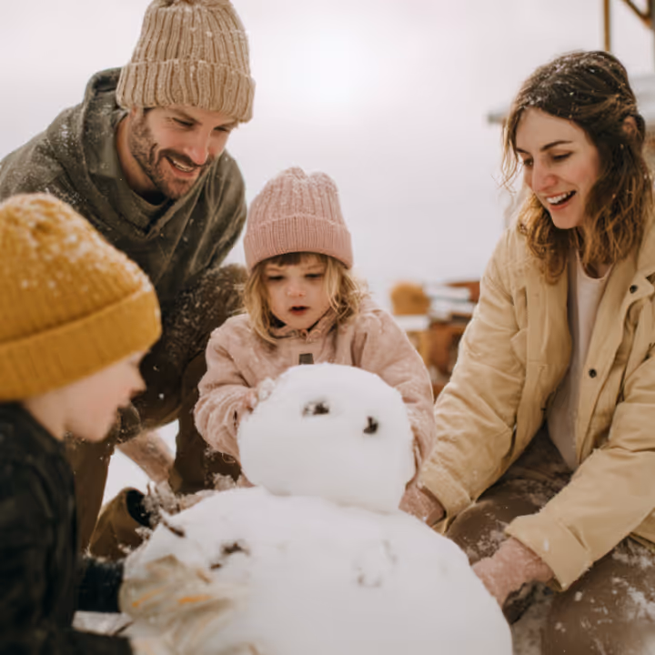 Family of four wearing winter clothes building a snowman indoors.
