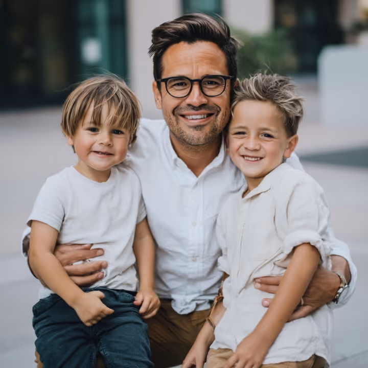 Smiling man wearing glasses hugging two young boys outdoors.
