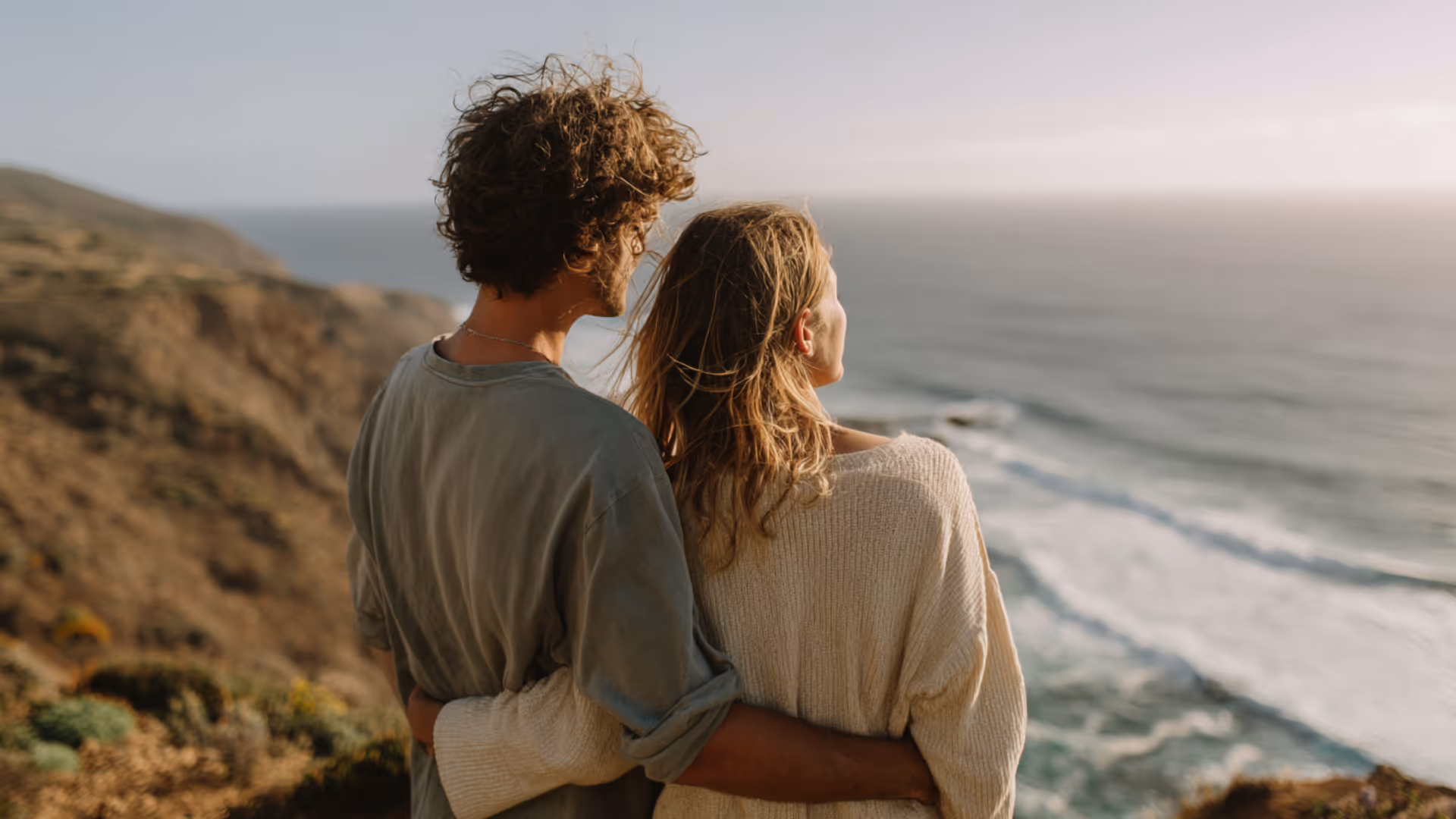 Couple embracing and looking out towards the ocean from a cliff at sunset.