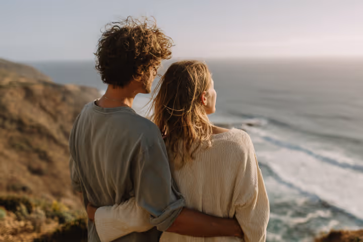 Couple embracing and looking out over ocean from a cliff during sunset.
