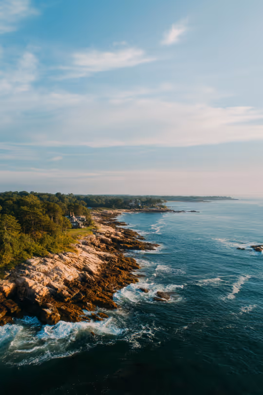 Aerial view of a rocky coastline with trees, houses, and waves crashing against the shore under a partly cloudy sky.