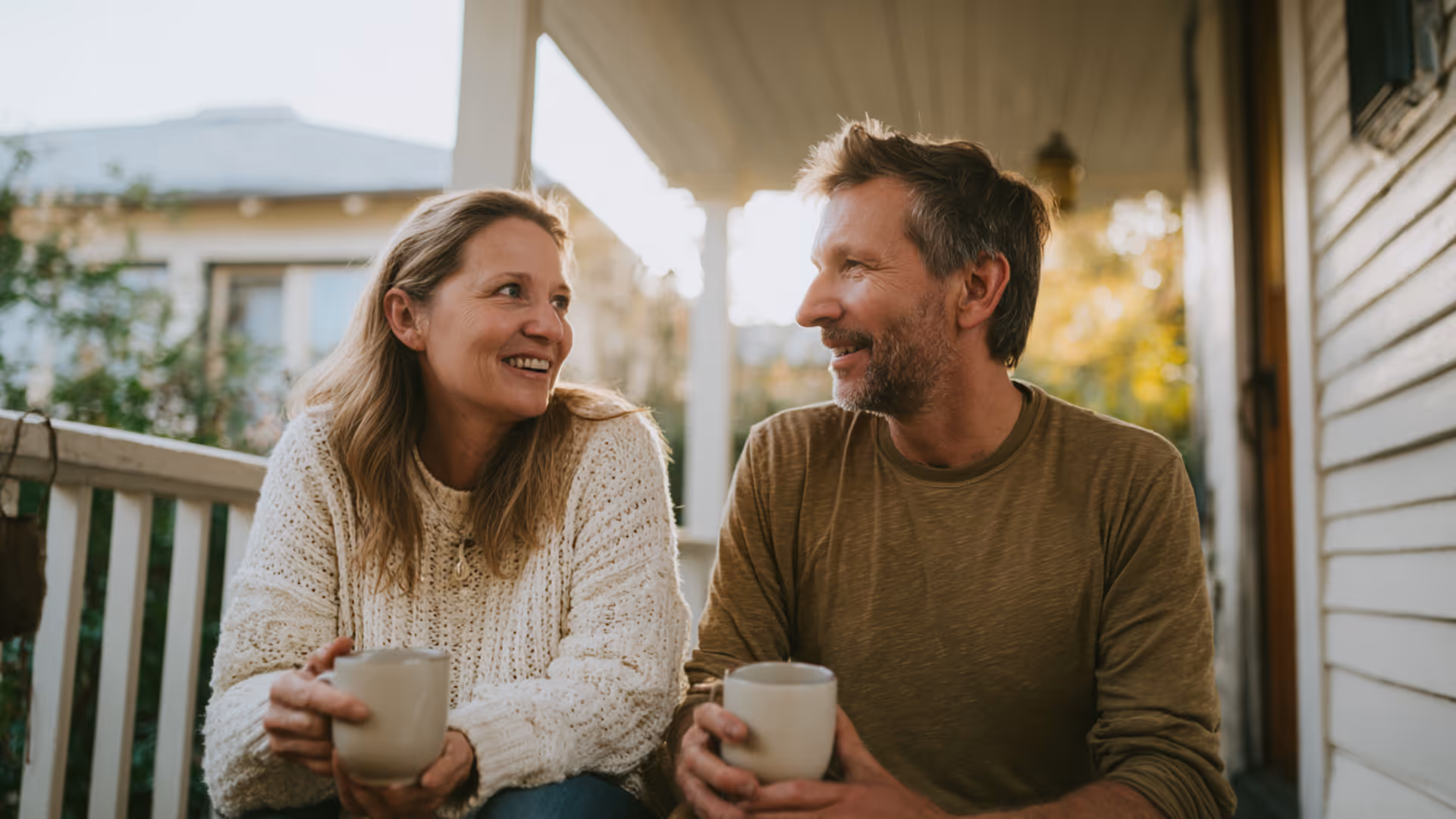 Smiling middle-aged couple sitting on a porch holding mugs and looking at each other.