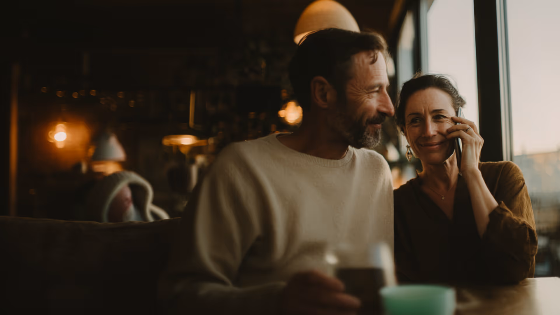 Smiling couple sitting close together in a cozy dimly lit cafe, with the woman holding a phone to her ear.