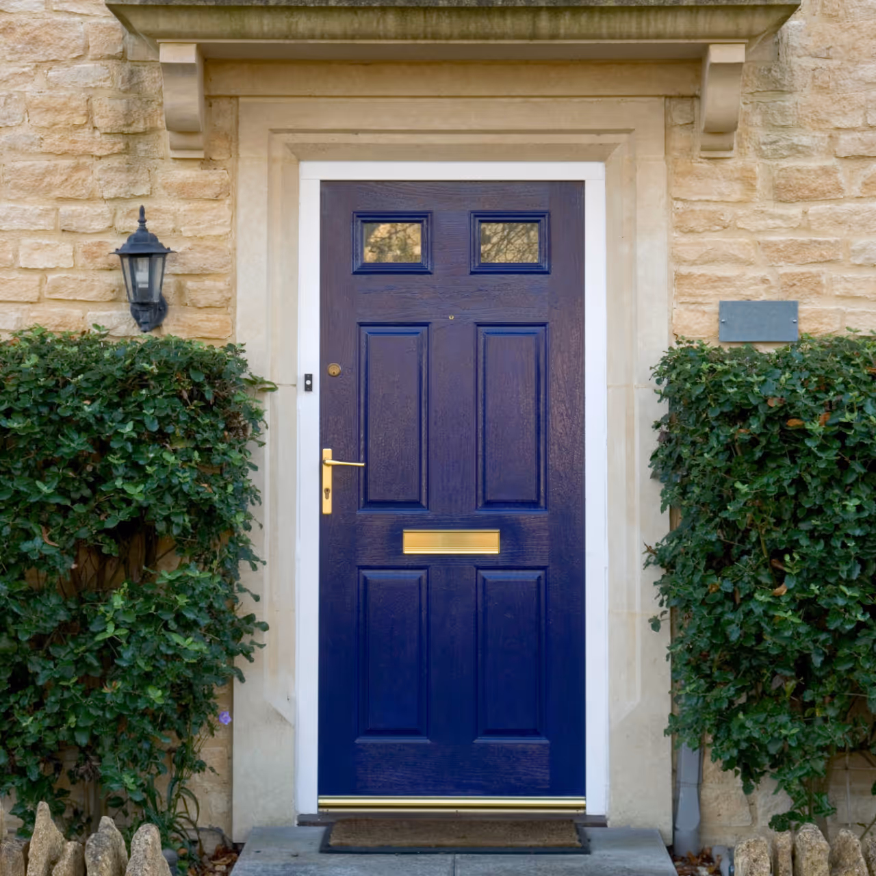 Blue front door with gold handle, mail slot, and door knocker set in a stone wall flanked by green bushes.