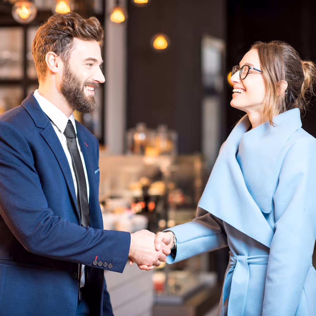 Smiling man in a blue suit and woman in a light blue coat shaking hands indoors.
