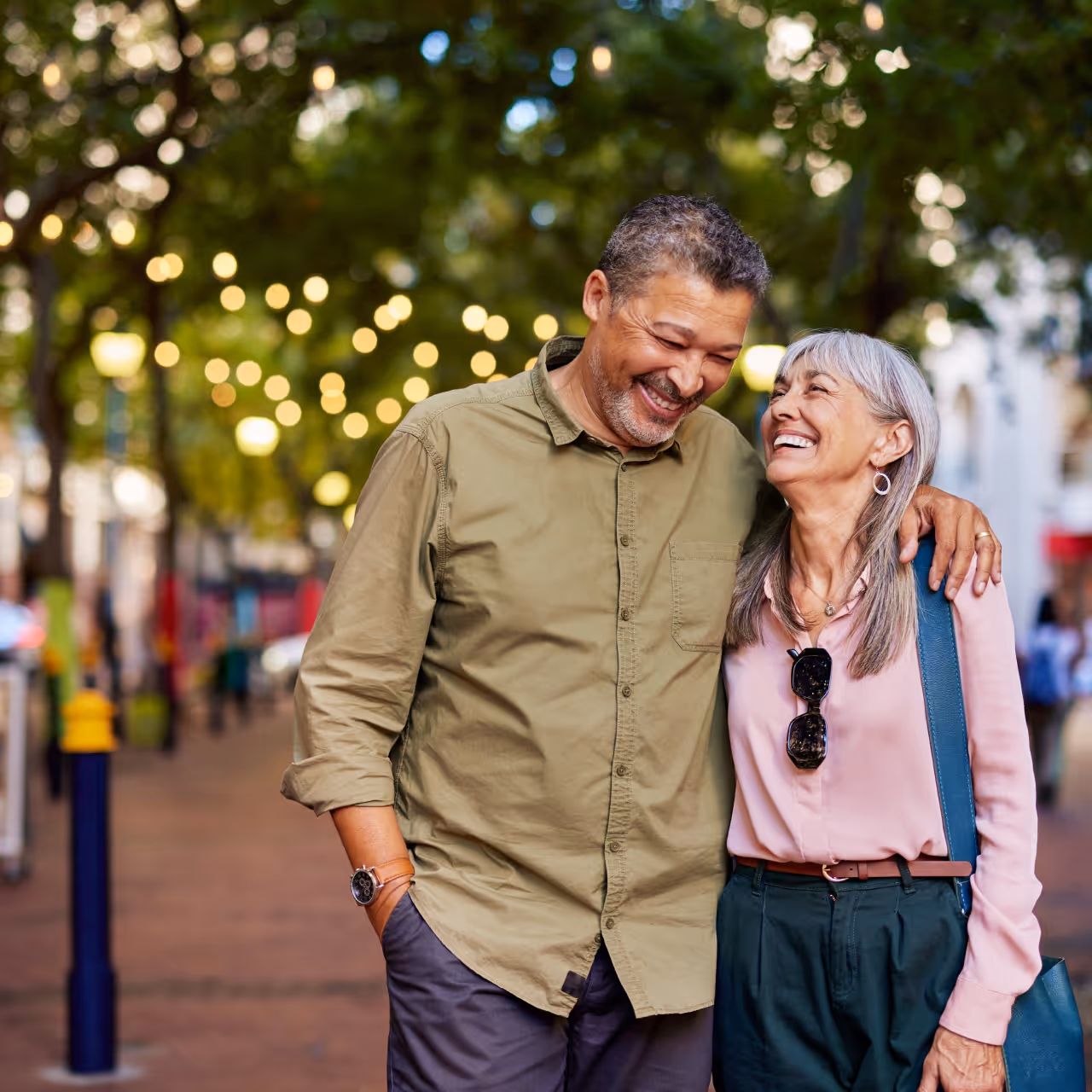 Smiling middle-aged couple walking arm in arm on a tree-lined street with blurred lights in the background.