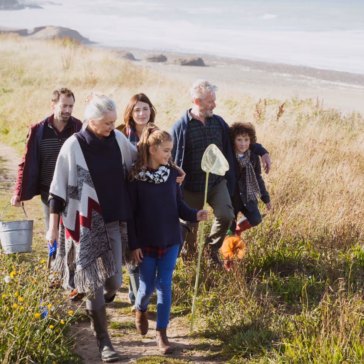 Multi-generational family walking outdoors on a grassy path near a beach, with a girl holding a butterfly net.