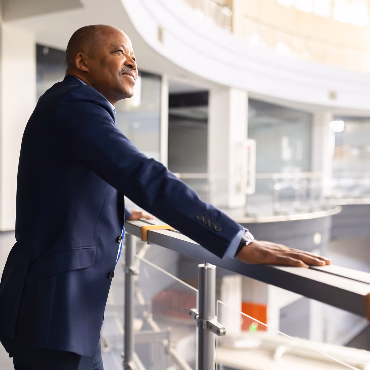 Confident senior businessman in navy suit leaning on balcony railing inside modern office building.