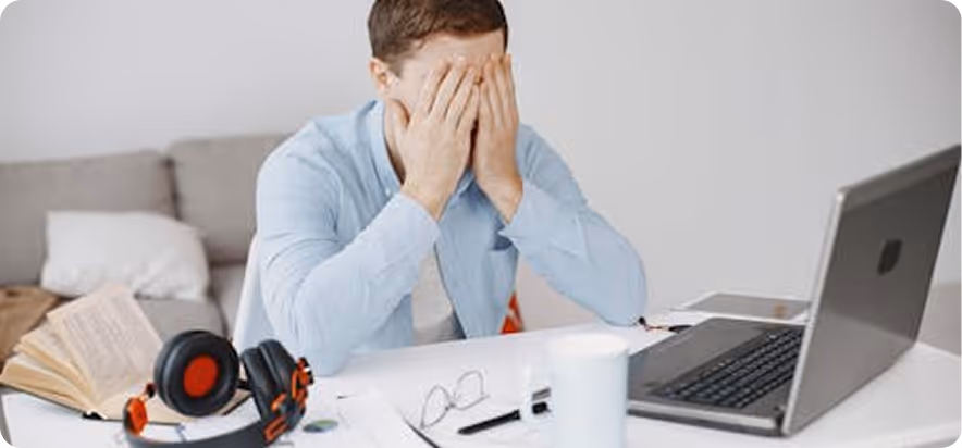 Man in blue shirt sitting at desk with laptop and headphones, covering his face with his hands.