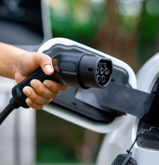 Ruka drži priključak za punjenje električnog vozila pored priključnog mesta na belom automobilu.