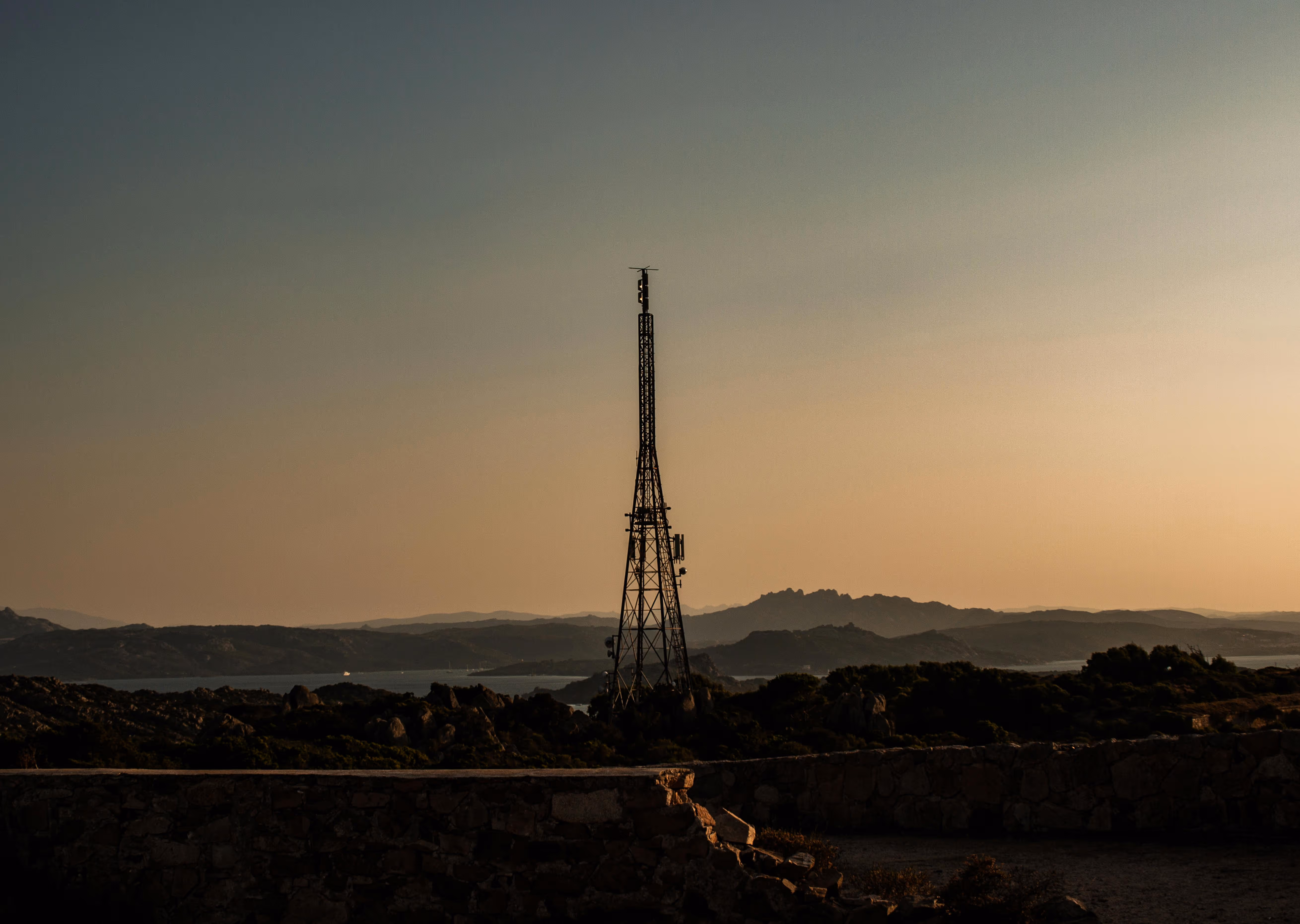 Metal antenna tower on rocky terrain with mountains and water under sunset sky.