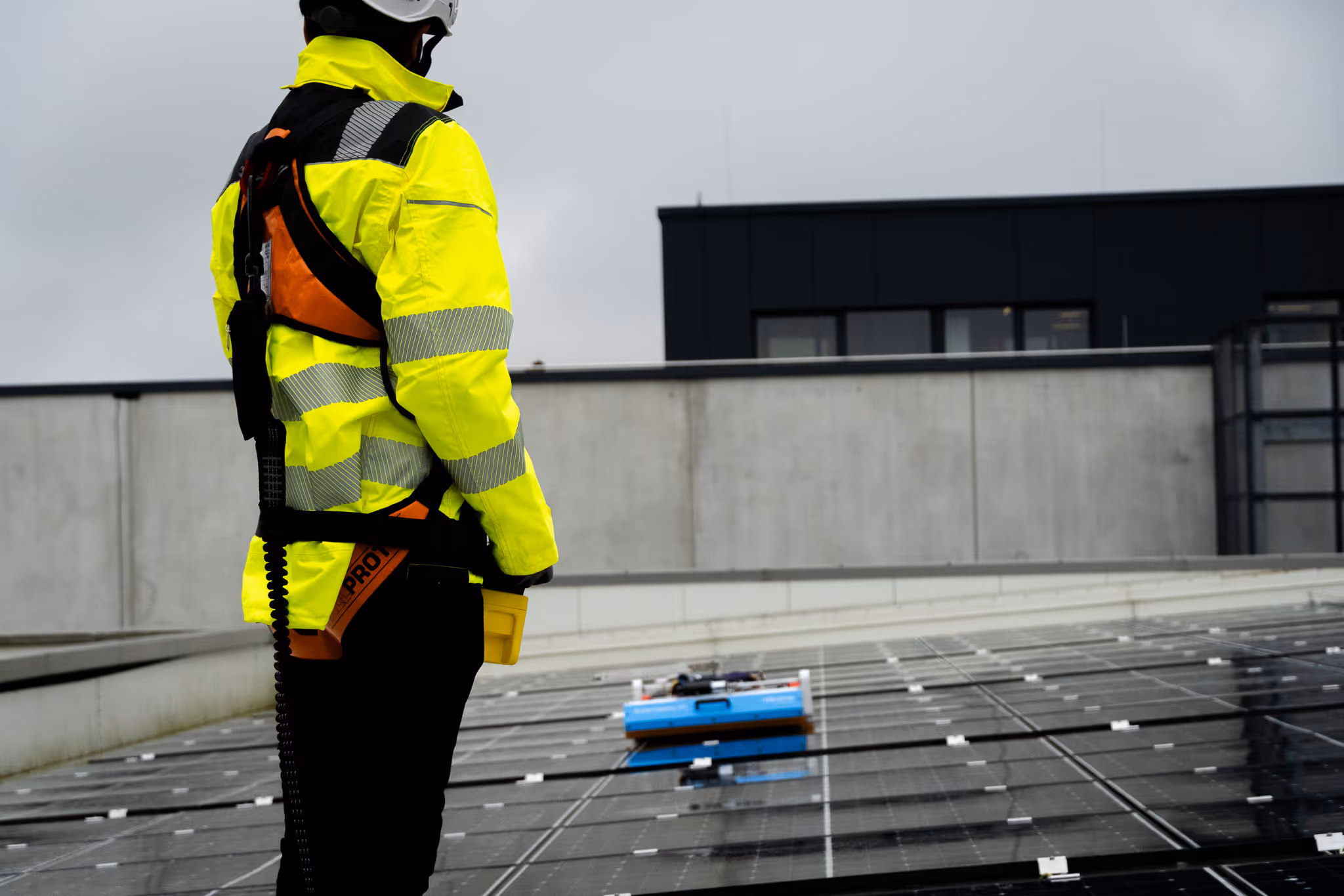 Worker in yellow safety jacket and helmet standing beside solar panels on a rooftop under cloudy sky.