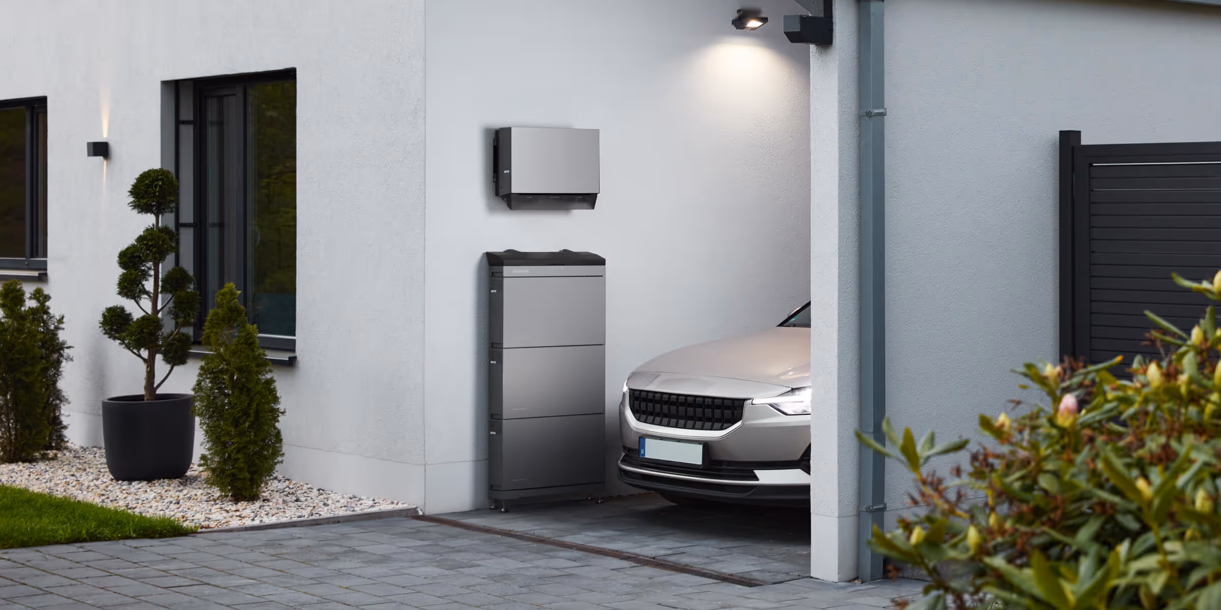 Silver car parked partially inside a modern garage with potted plants and a wall-mounted device.