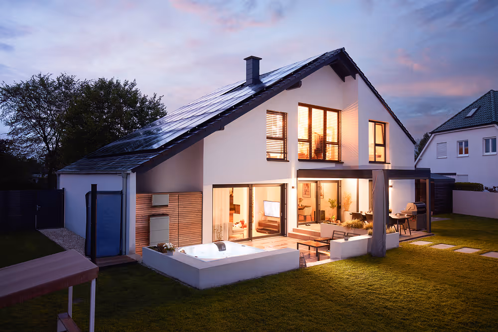 Modern two-story house with solar panels, large windows, outdoor hot tub, and lit interior at dusk.