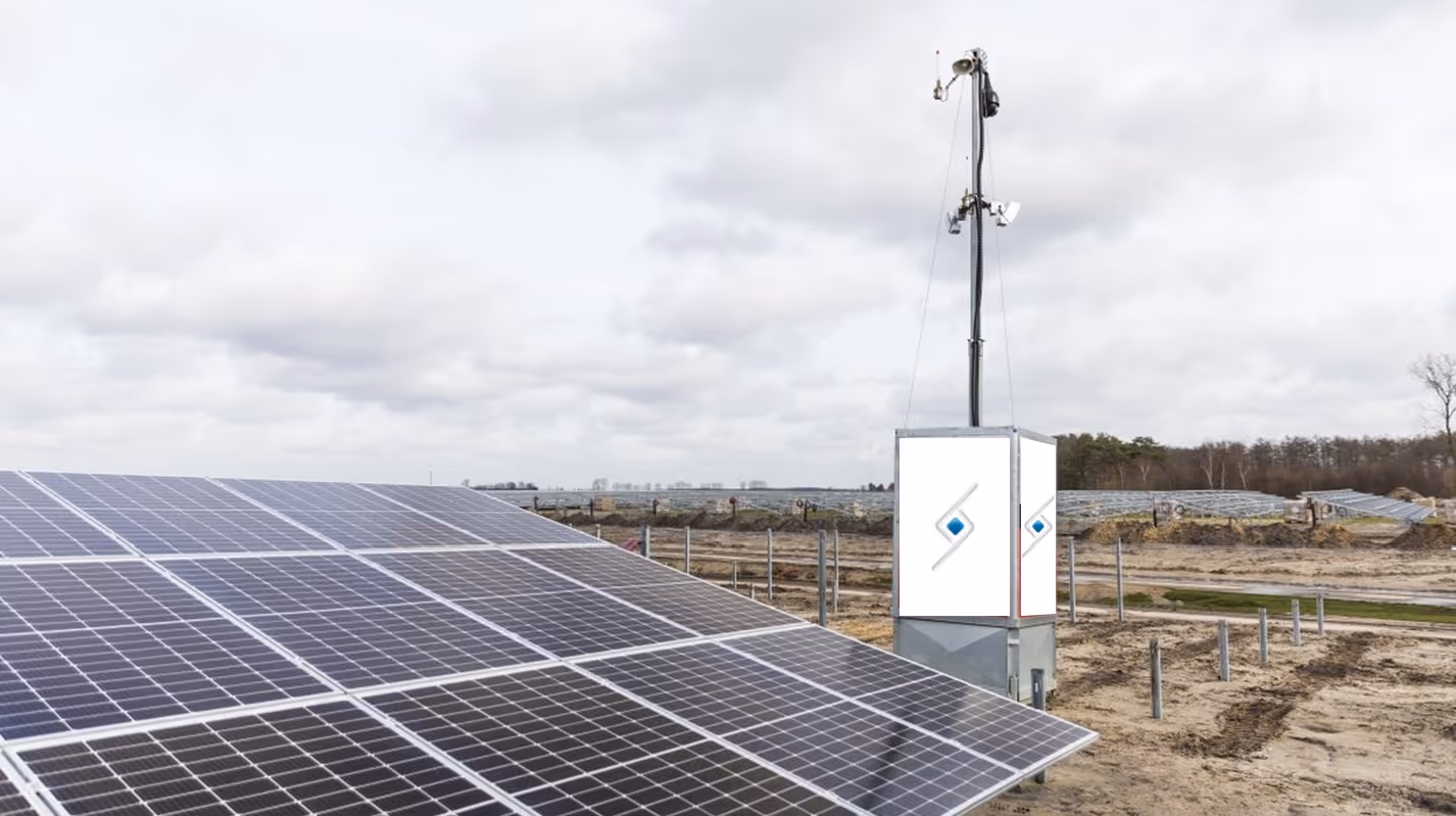 Solar panels in a field with a monitoring device mounted on a pole under a cloudy sky.