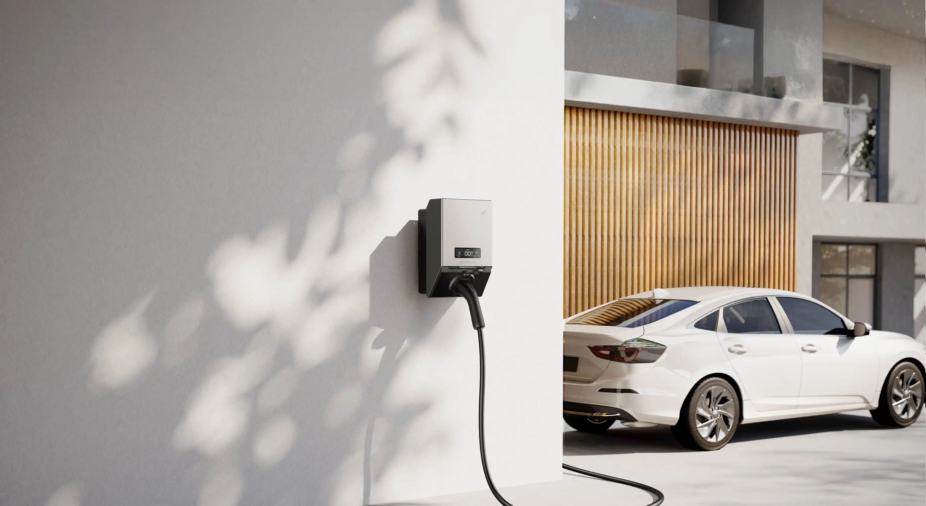 Electric vehicle charger mounted on a white wall with a white car parked nearby in front of a wooden garage door.