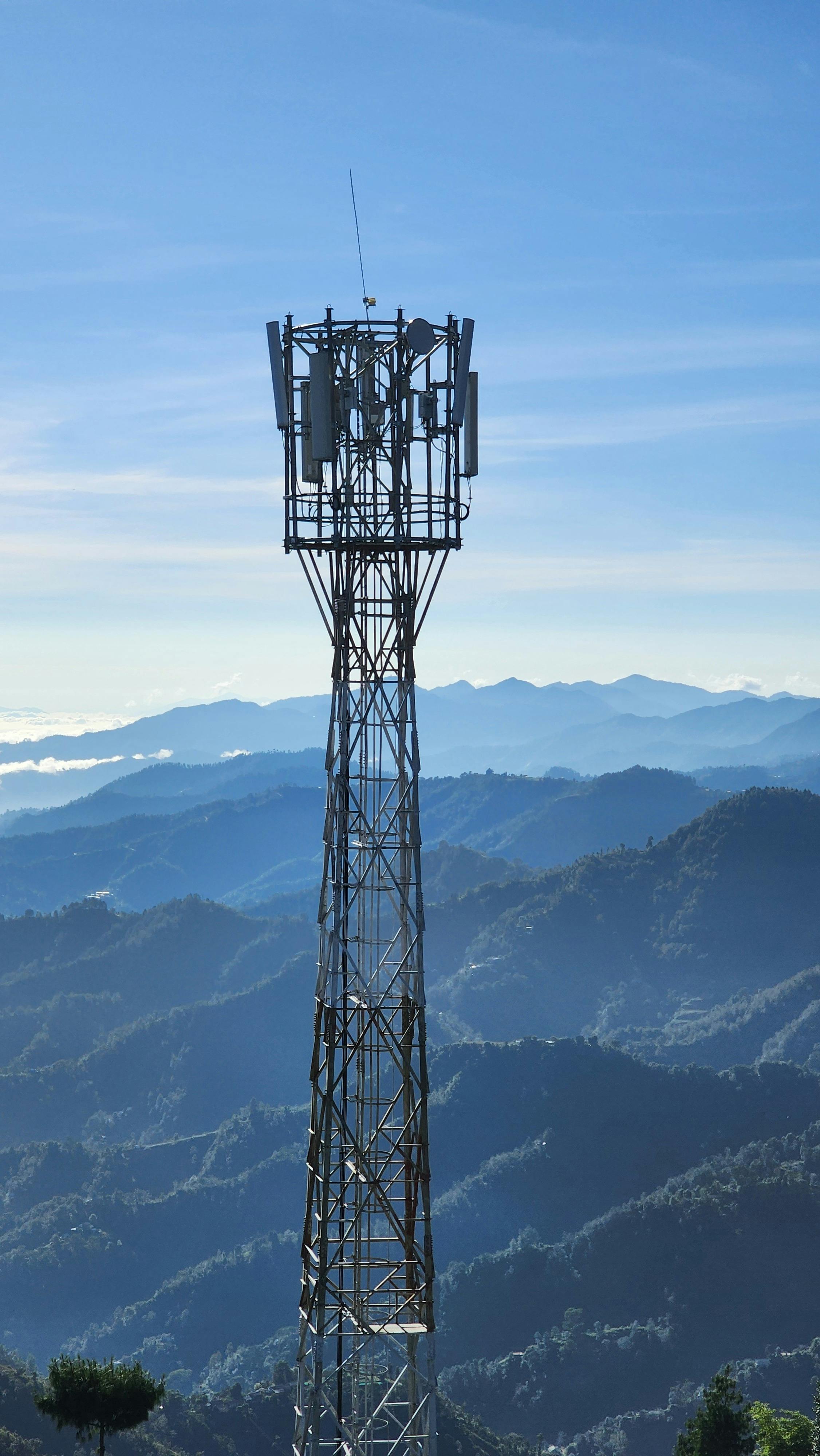 Metal communication tower with antennas in front of layered blue mountain ranges under a clear sky.