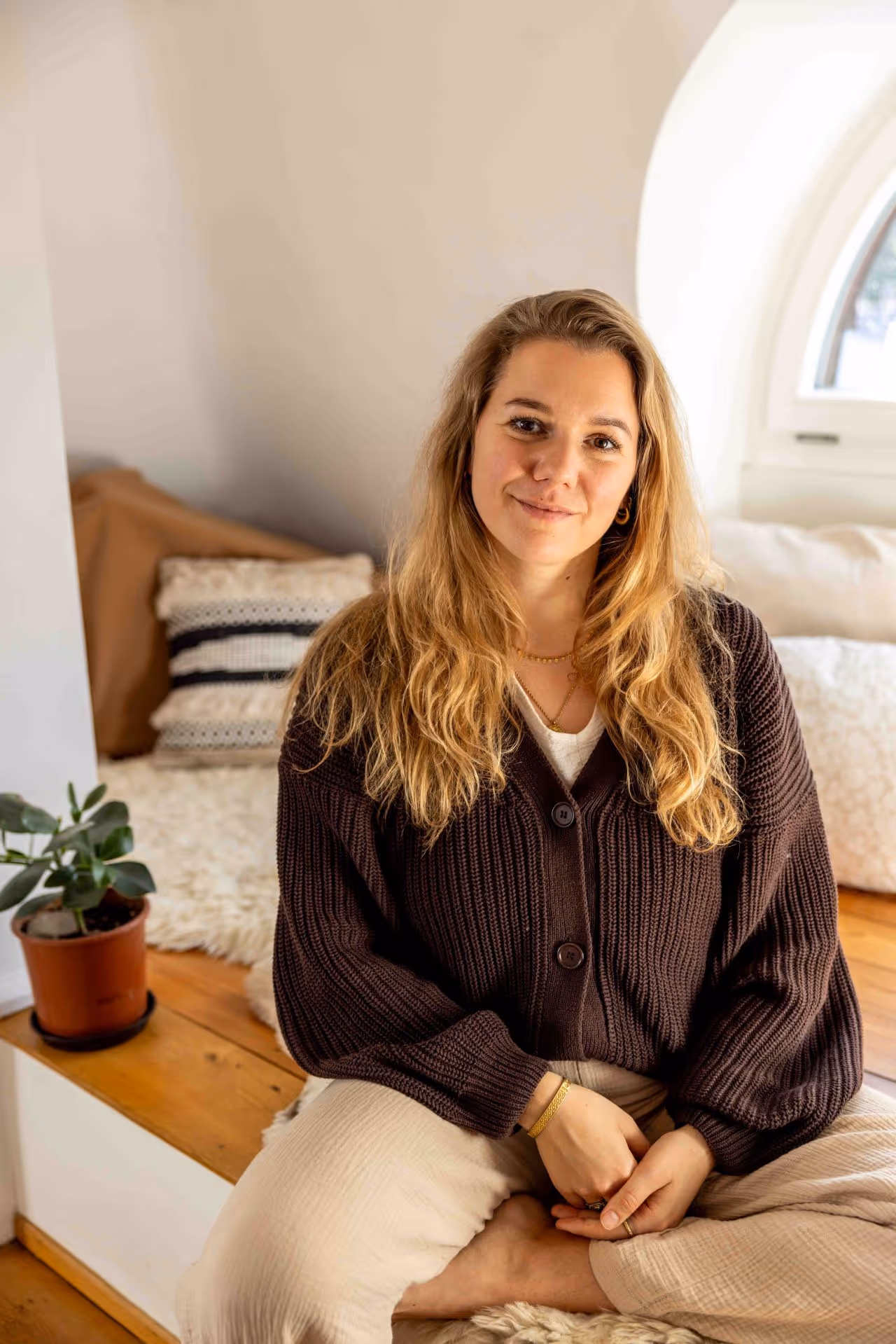 Junge Frau mit blonden Haaren sitzt entspannt in einem gemütlichen Raum mit Kissen und Pflanzen.