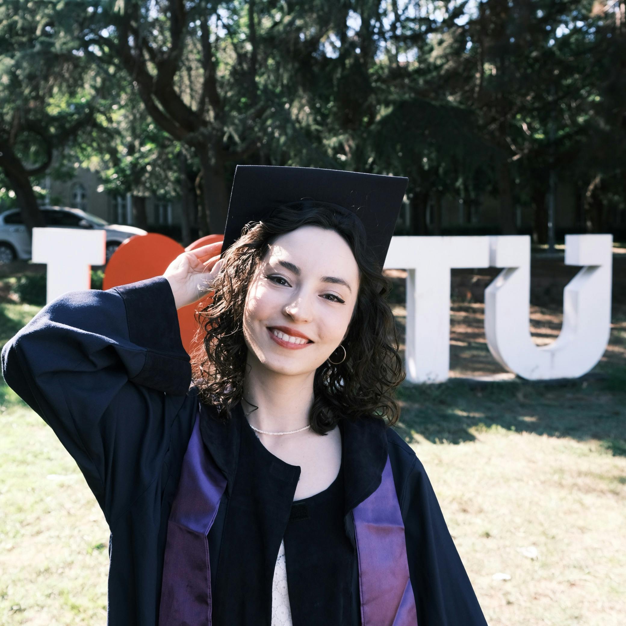 Smiling young woman in graduation gown and cap touching her hat outdoors with large letters and trees in the background.