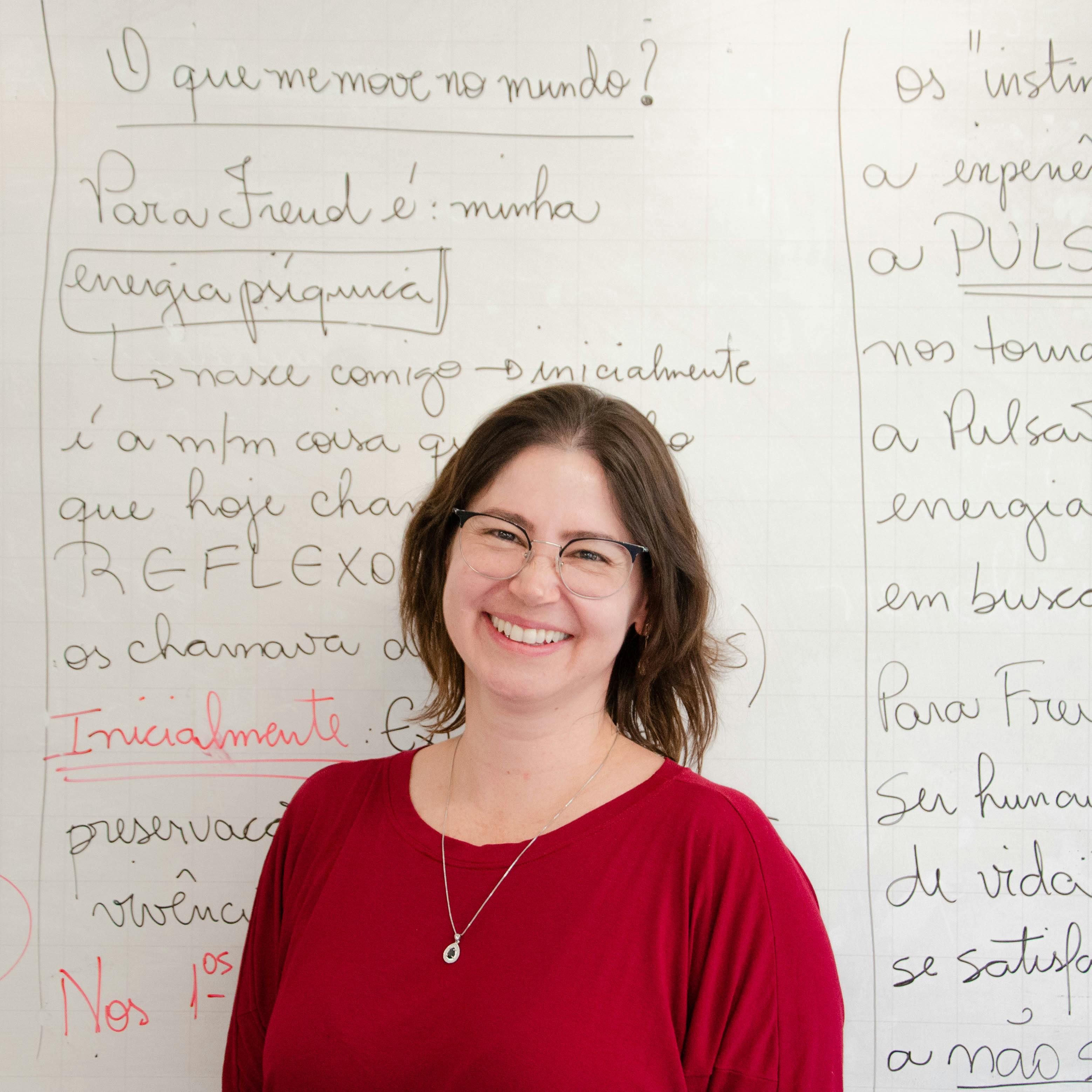 Smiling woman with glasses and a red top standing in front of a whiteboard filled with handwritten text in Portuguese.