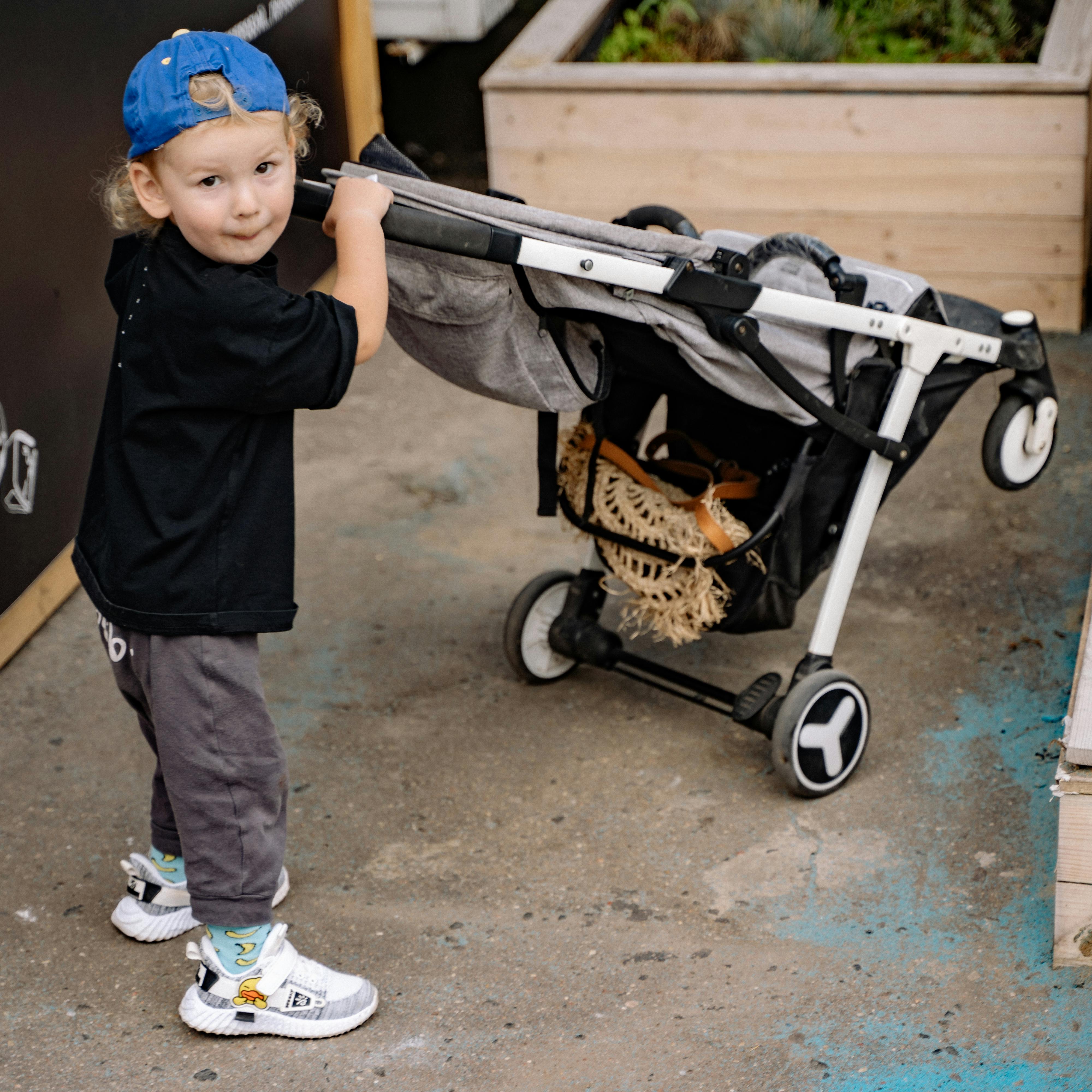Young child wearing a blue backward cap and black shirt pushing a gray stroller on a concrete surface.