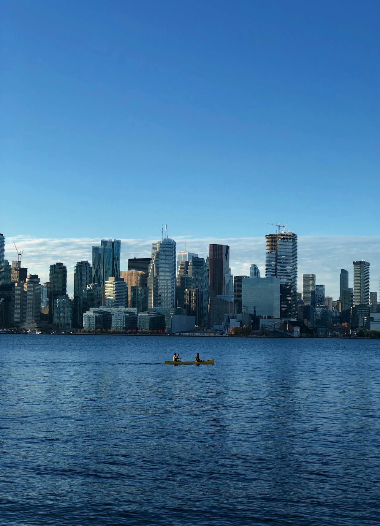 Two people paddling a yellow canoe on water with a modern city skyline in the background under a clear blue sky.