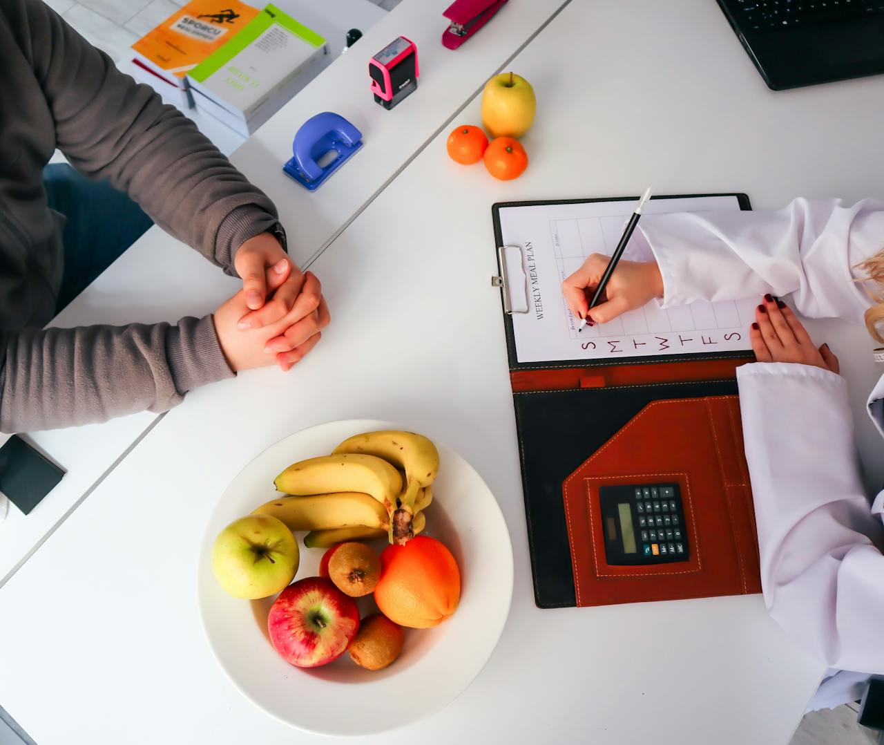 Person in a white coat writing a weekly meal plan on a clipboard across from another person with folded hands at a table with fruits and office supplies.