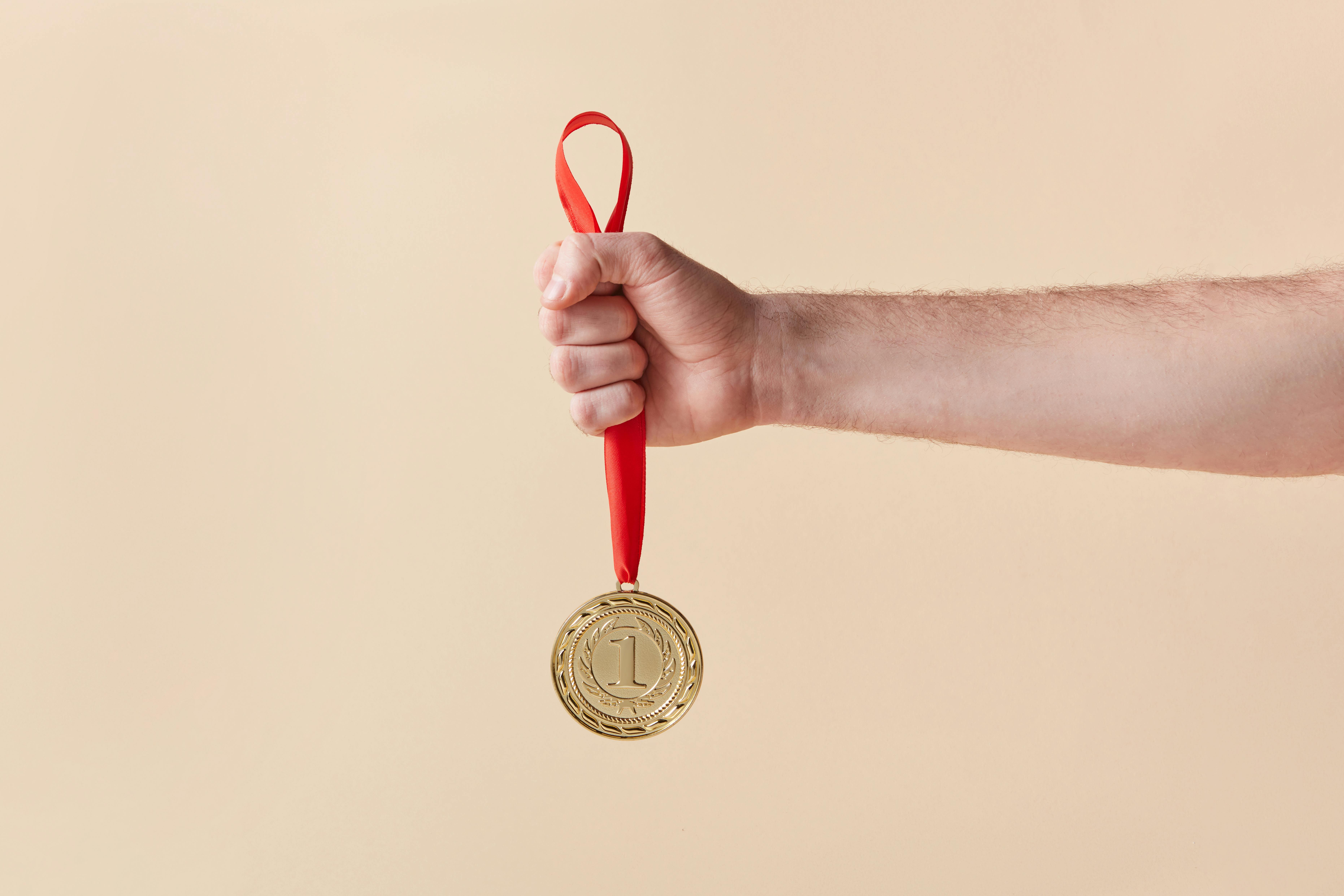 Hand holding a gold first place medal with a red ribbon against a beige background.