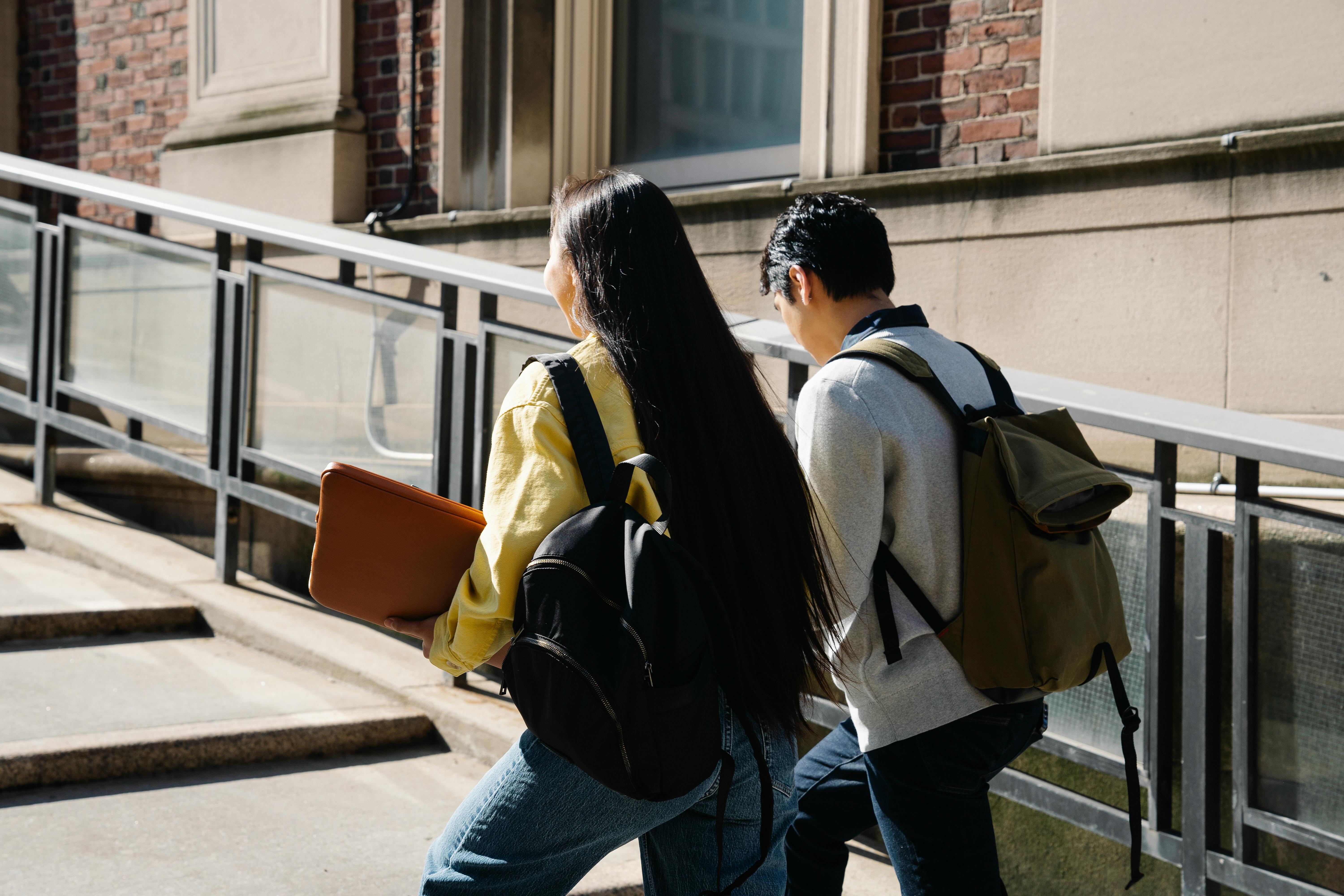 Two students walking up outdoor steps beside a building, one carrying an orange folder and wearing a black backpack, the other with a green backpack.
