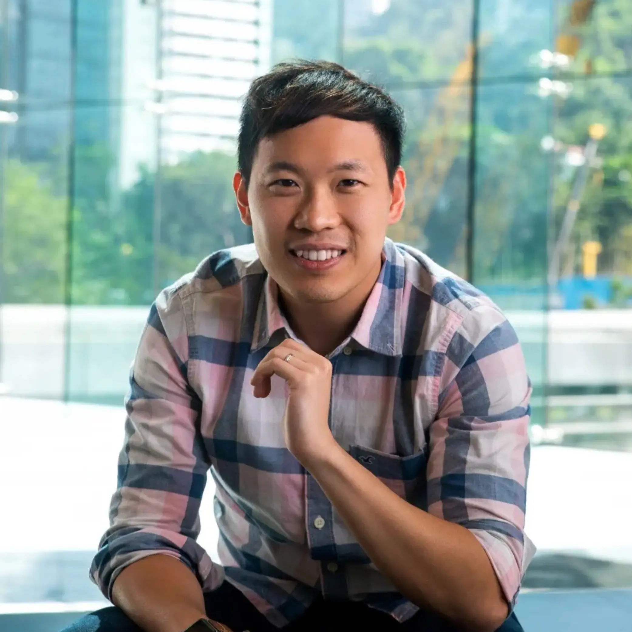 Smiling man in a plaid shirt sitting indoors with a glass window and city view behind him. education marketing agency WonderMaple Strategy