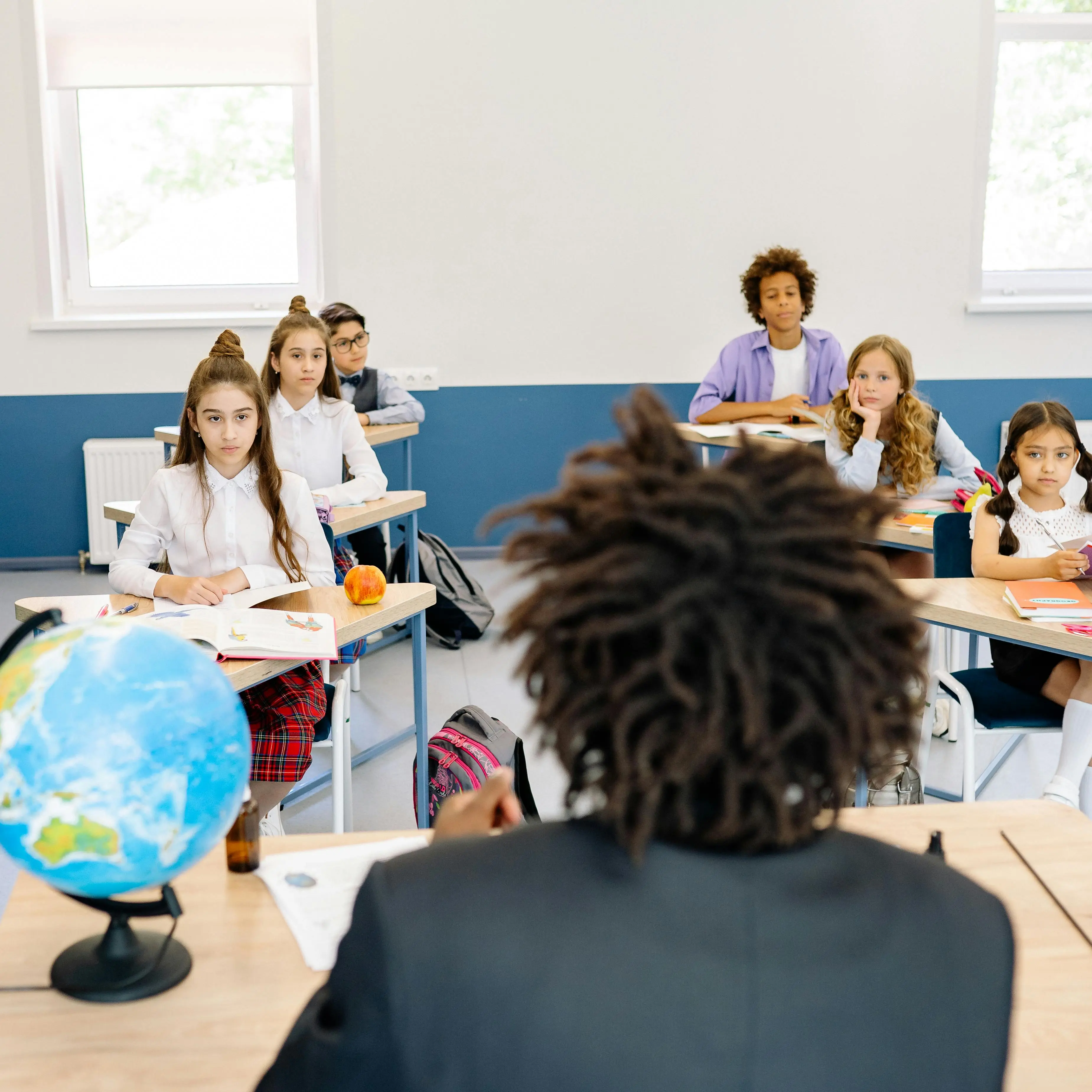 Teacher with dreadlocks facing a classroom of attentive students sitting at desks.