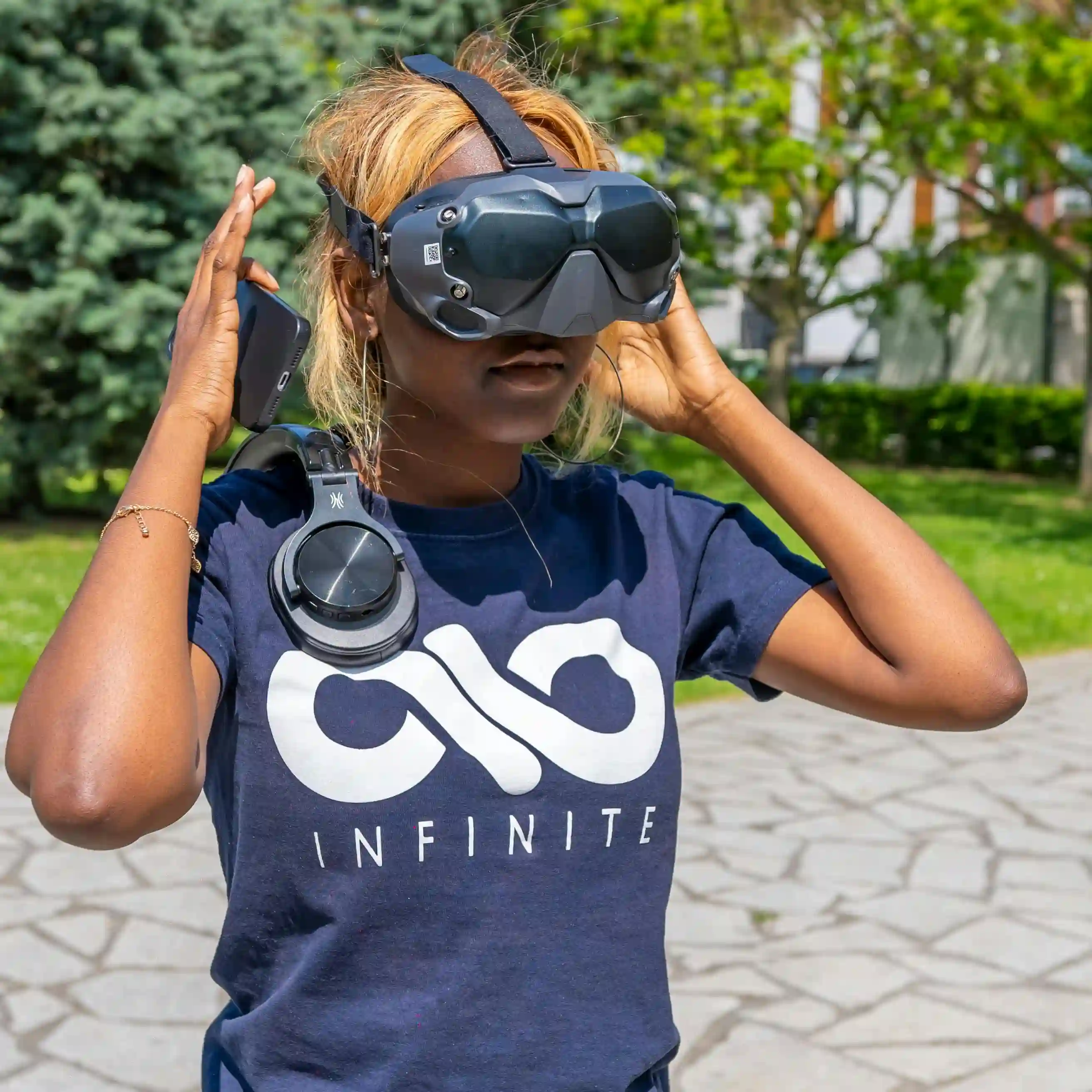 Person outdoors wearing a VR headset and a navy blue T-shirt with the word INFINITE, holding a phone and black headphones around their neck.