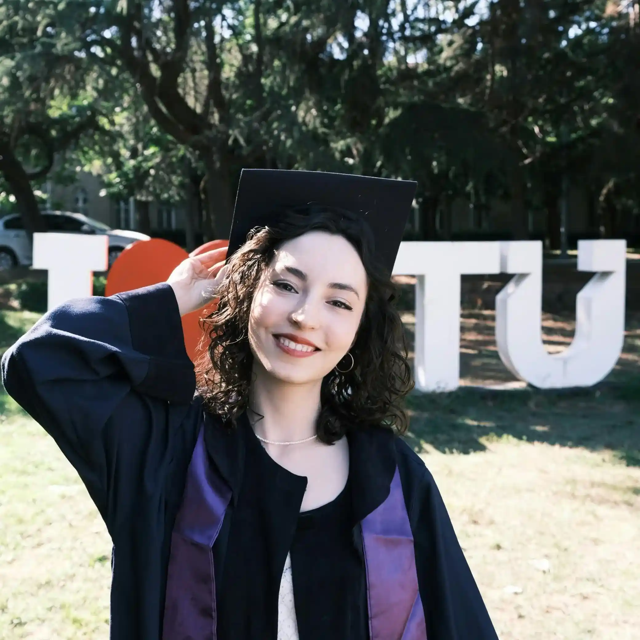 Smiling young woman in graduation gown and cap touching her hat outdoors with large letters and trees in the background.
