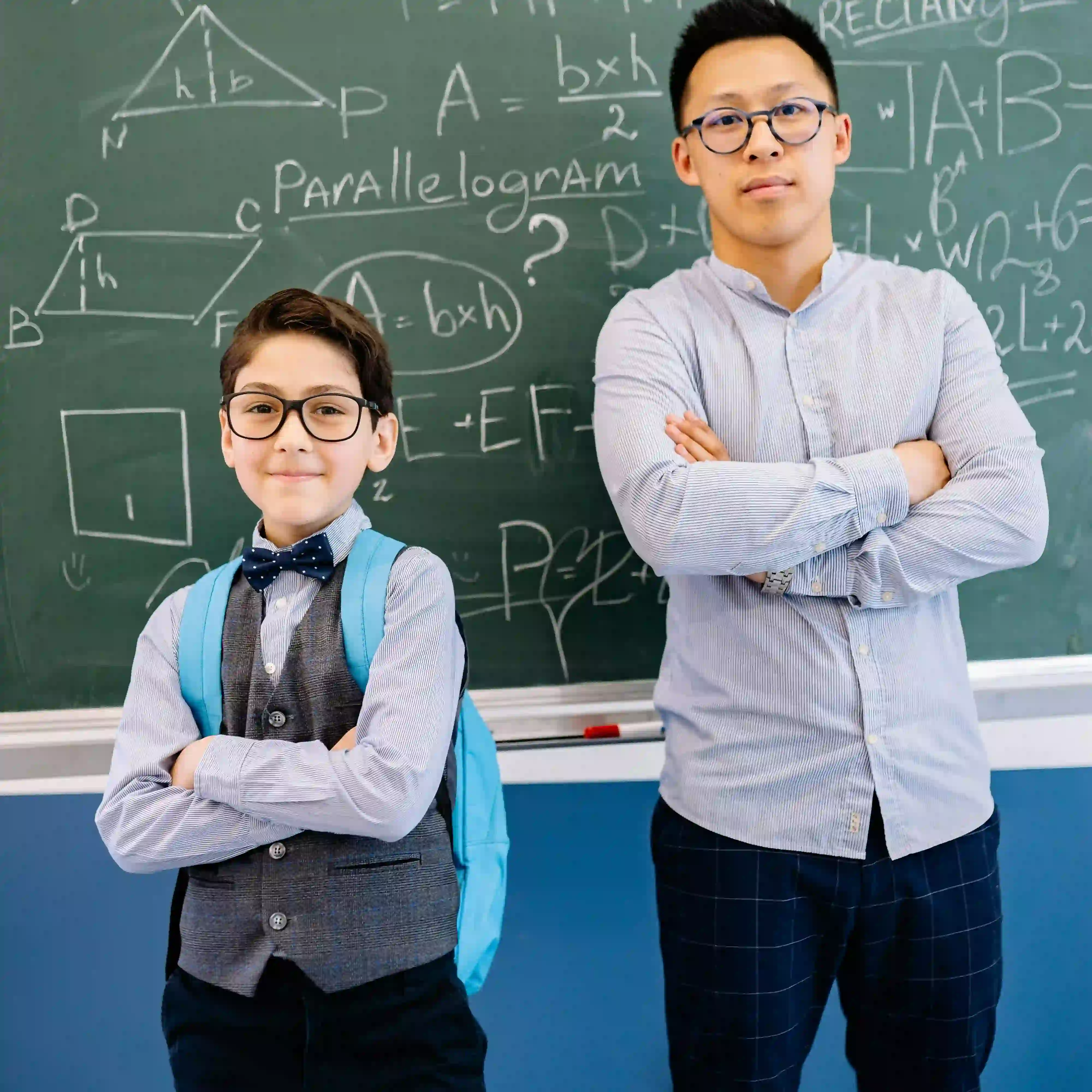 Young boy with glasses and backpack standing confidently next to a teacher in front of a chalkboard with math formulas.